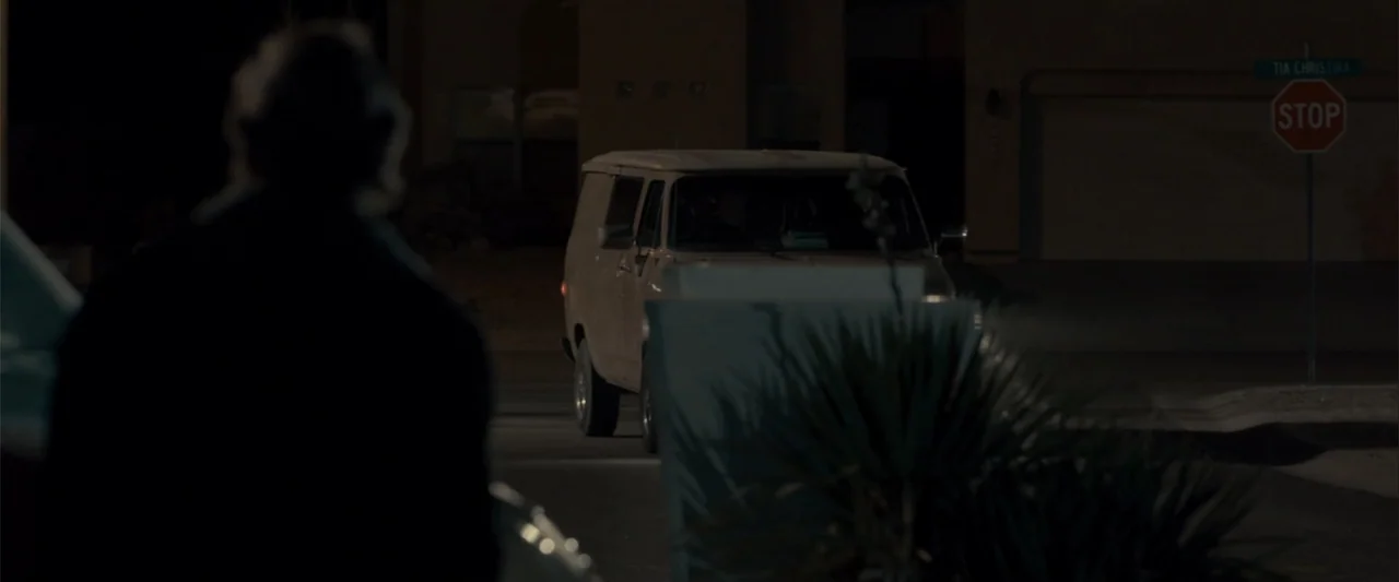 Frontera (2014) movie still: medium — A dark residential street corner features a parked vintage van under low ambient lighting. A blurry figure…