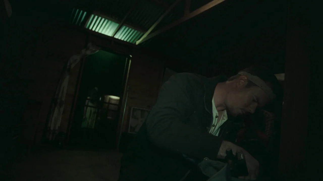 The Sacrament (2013) movie still: medium — A dark, cramped wooden room features corrugated metal roofing and a doorway illuminated green. A man…