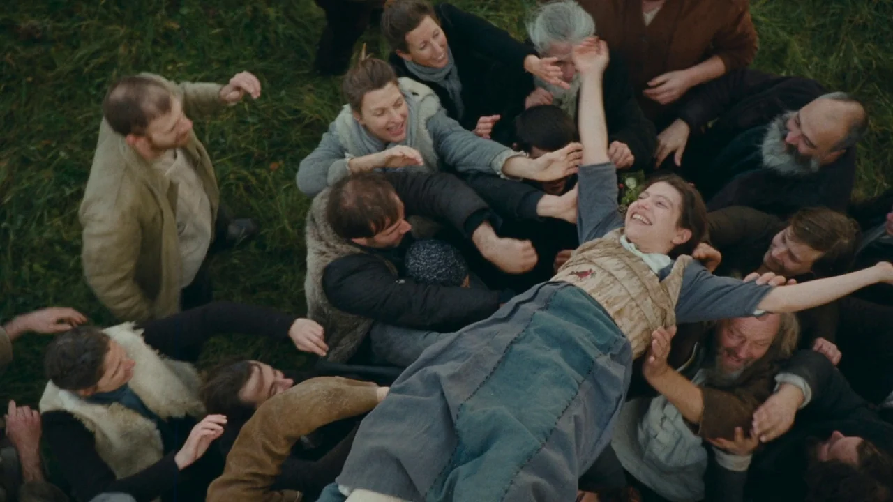 The Devil’s Bath (2024) movie still: wide — A group of people wearing simple rustic clothing gathered together in a large field. A young woman is…