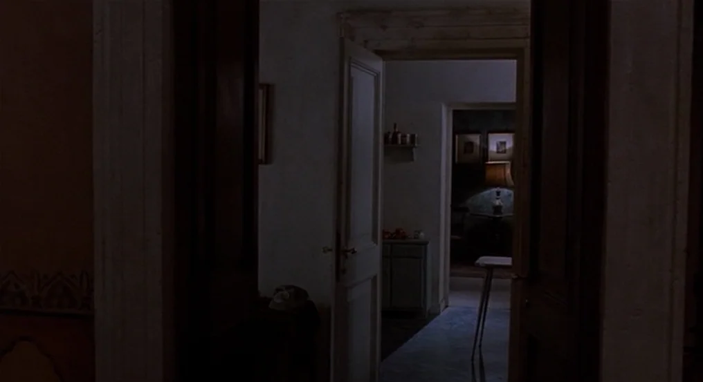 The Comfort of Strangers (1990) movie still: wide — A dimly lit residential apartment hallway connects multiple rooms through open doorways. Dark…