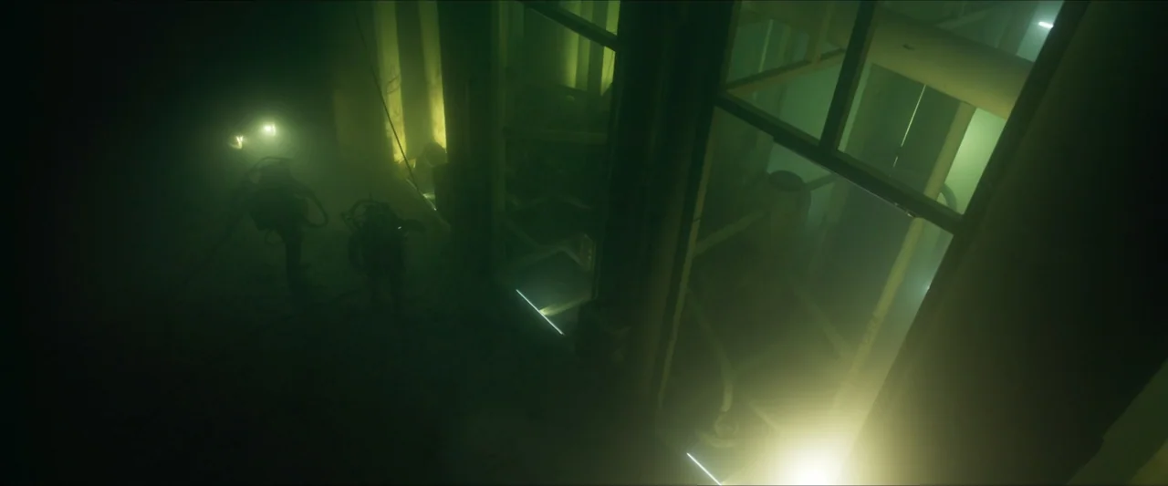 Last Breath (2025) movie still: wide — A dimly lit industrial interior filled with heavy fog and green ambient lighting. Two figures wearing scuba…
