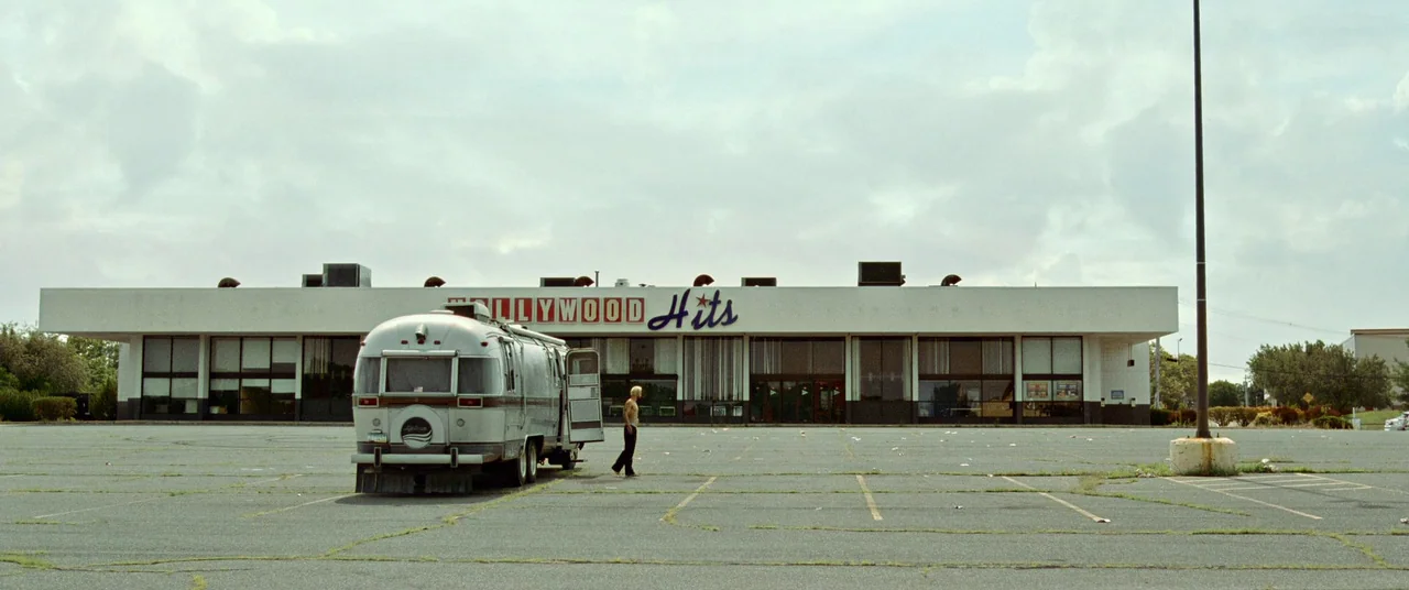 Sound of Metal (2019) movie still: wide — A wide, empty parking lot features a large commercial building under an overcast sky. A silver…