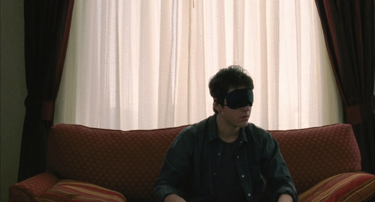 Incendies movie still: medium — A man sits on a reddish-patterned sofa in front of large, floor-to-ceiling sheer curtains. The man wears a black…