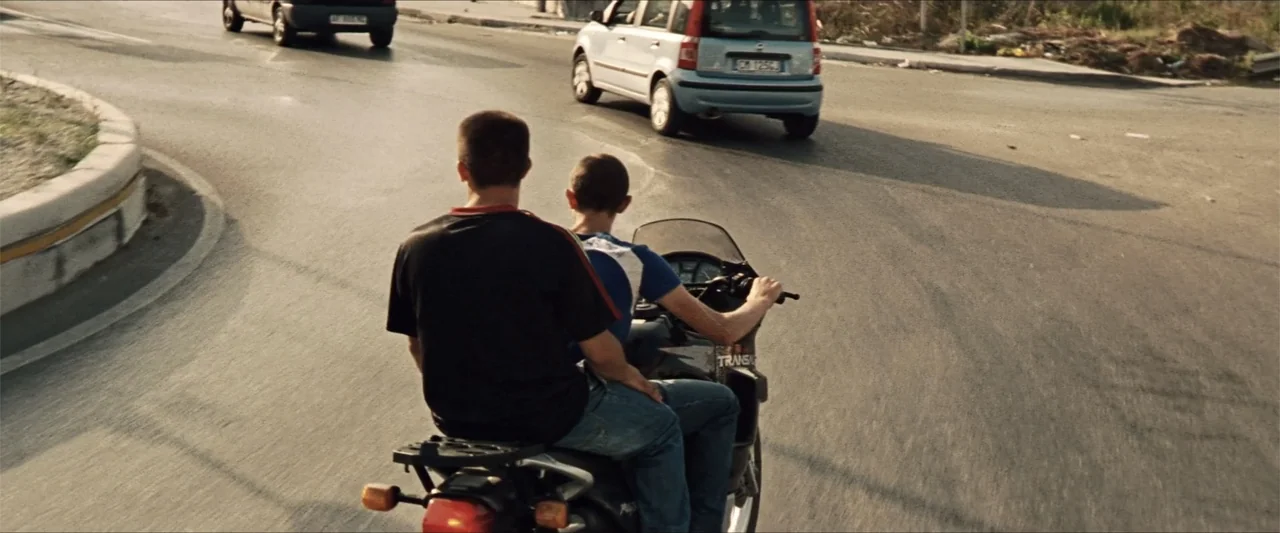 GOMORRAH (2023) movie still: medium — An asphalt road intersection features road markings and passing light blue and black cars. Two males sit on a…