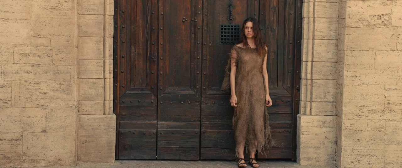 Benedetta (2021) movie still: fullbody — A woman stands against a massive, dark wooden door set within weathered stone walls. She wears a tattered,…