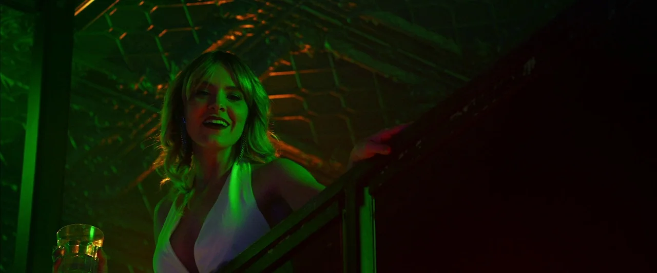 The Stylist (2020) movie still: medium — A dark balcony overlooks a dim club space with overhead patterned metal ceiling tiles. A woman wearing a…