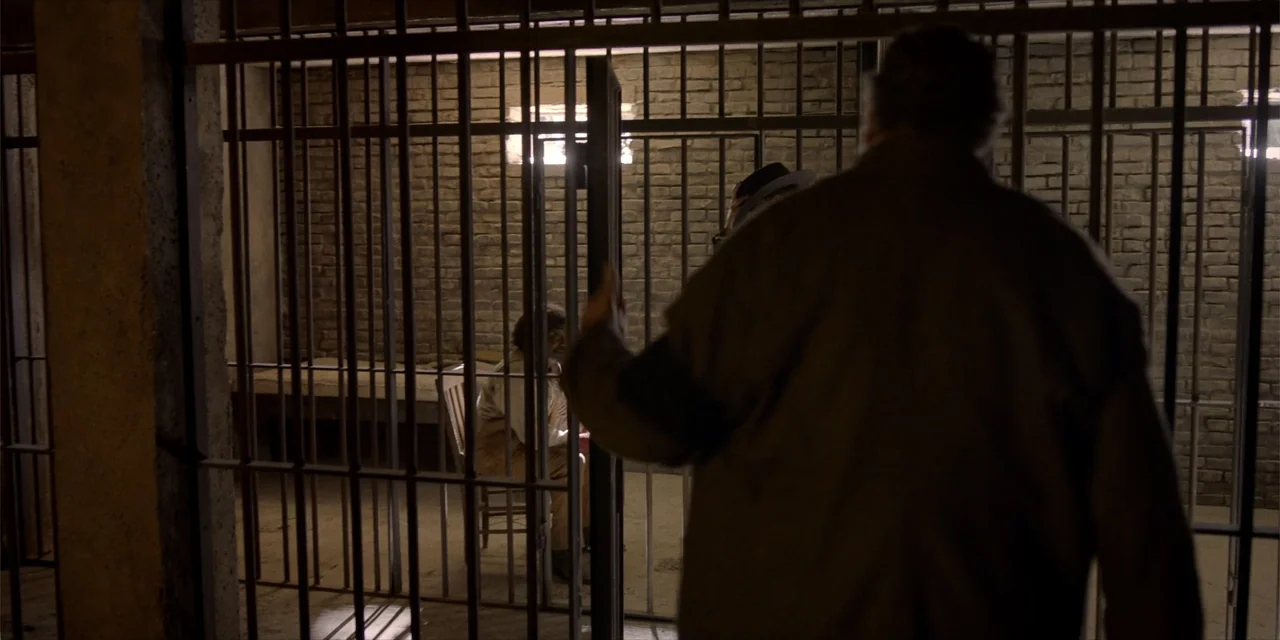 Deadwood: The Movie (2019) movie still: medium — A dimly lit stone enclosure features vertical metal bars and a plain wooden chair. A man standing…