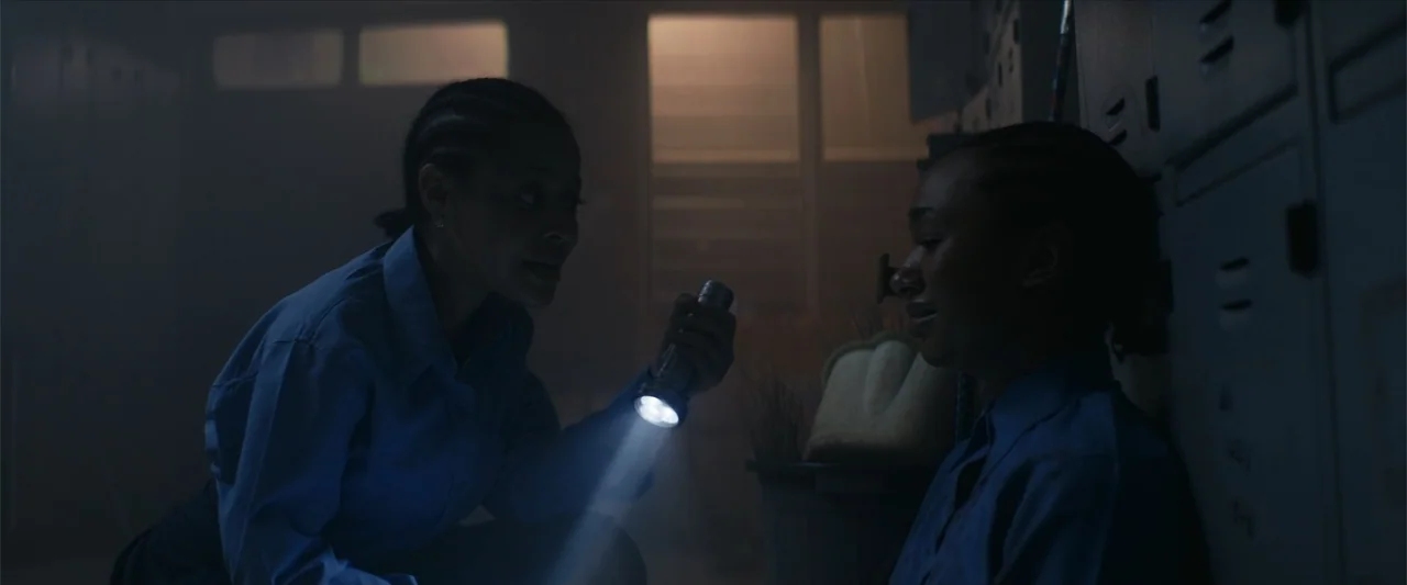 Ms. Kanyin (2025) movie still: medium — A dimly lit locker room features rows of metal lockers and visible dust particles. One woman shines a…