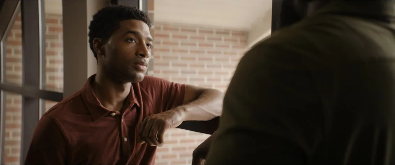 The Forge (2024) movie still: medium — An office interior features a brick wall background visible through a glass partition. A young man in a…