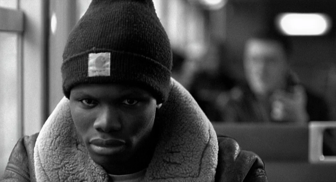 La Haine (1995) movie still: closeup — A person sits on a commuter train with blurred seating rows visible in the back. The individual wears a knit…