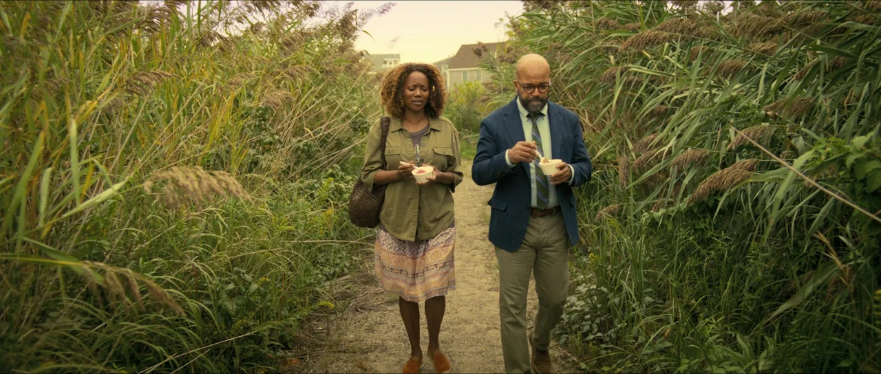 American Fiction (2023) movie still: fullbody — A man and woman walk along a narrow dirt path flanked by tall marsh grasses. The duo carries small…