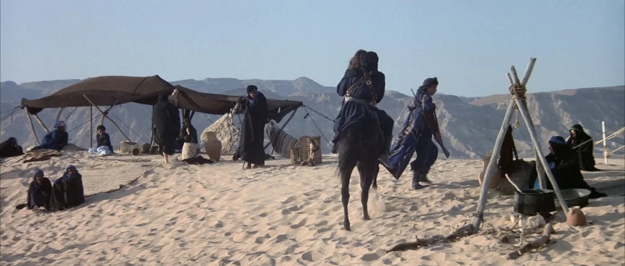 Sahara (1995) movie still: wide — A desert encampment with several canvas tents and small groups of people situated. Figures dressed in long dark…