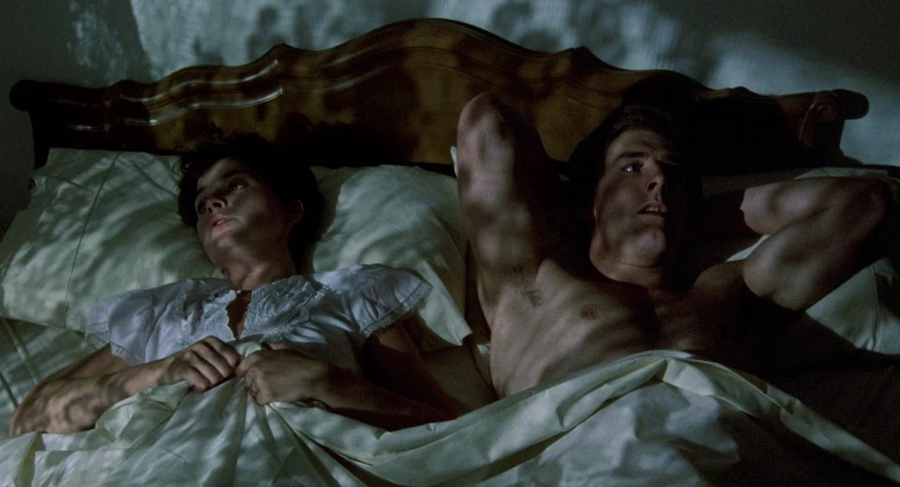 Crimes of Passion (1984) movie still: medium — A man and a woman lie side-by-side in a bed with a wooden headboard. They are awake and staring ahead…