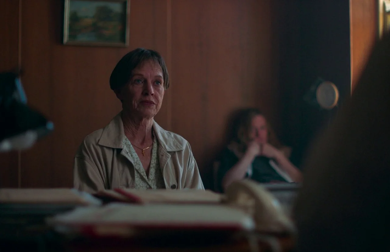 Nitram (2021) movie still: medium — A wood-paneled room features a desk cluttered with papers and a telephone in foreground. A mature woman sits at…