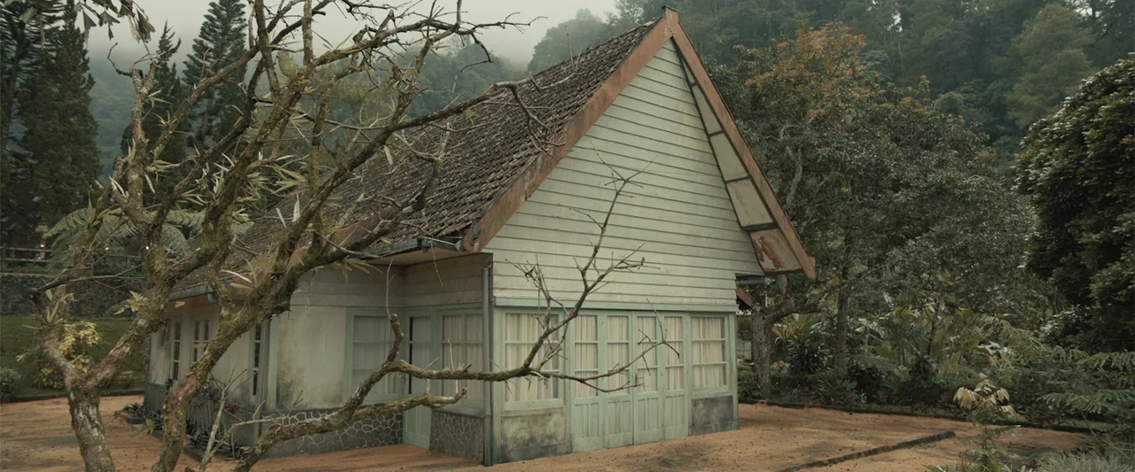 The Unholy (2022) movie still: establishing — A light-colored wooden cabin with a gable roof sits in a misty mountainous landscape. The surrounding…