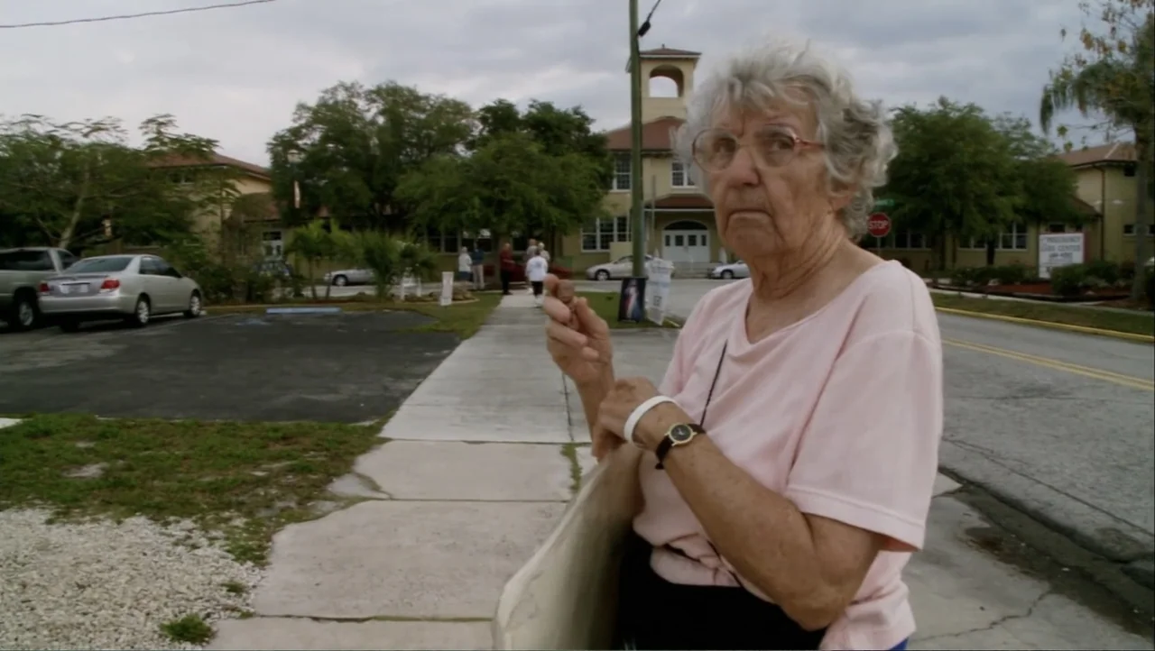 12th & Delaware (2010) movie still: medium — An outdoor sidewalk is located in front of a multi-story institutional brick building. An elderly woman…