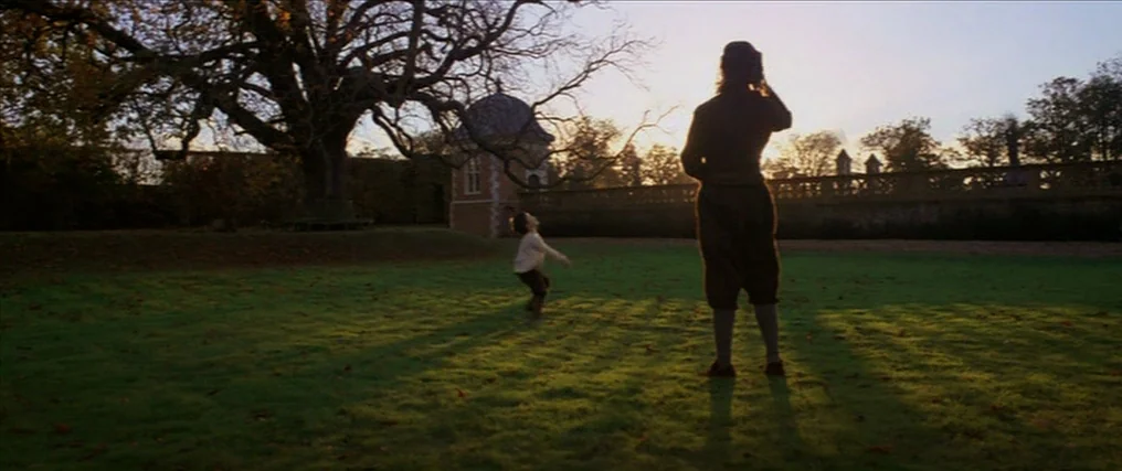 The New World (2005) movie still: wide — A sprawling estate lawn is dominated by a large leafless tree at sunset. A man stands in the foreground…