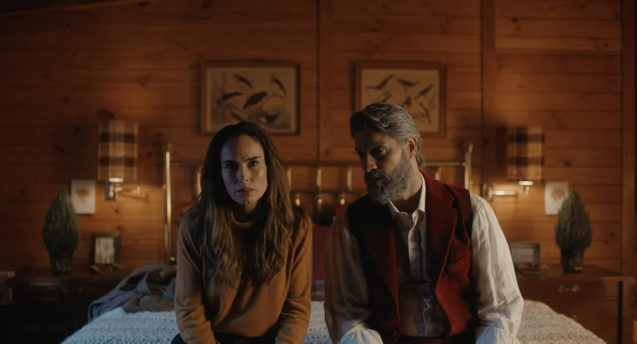Dating Santa (2023) movie still: medium — A room with wood-paneled walls decorated with framed bird sketches above a bed. A man and a woman sit…