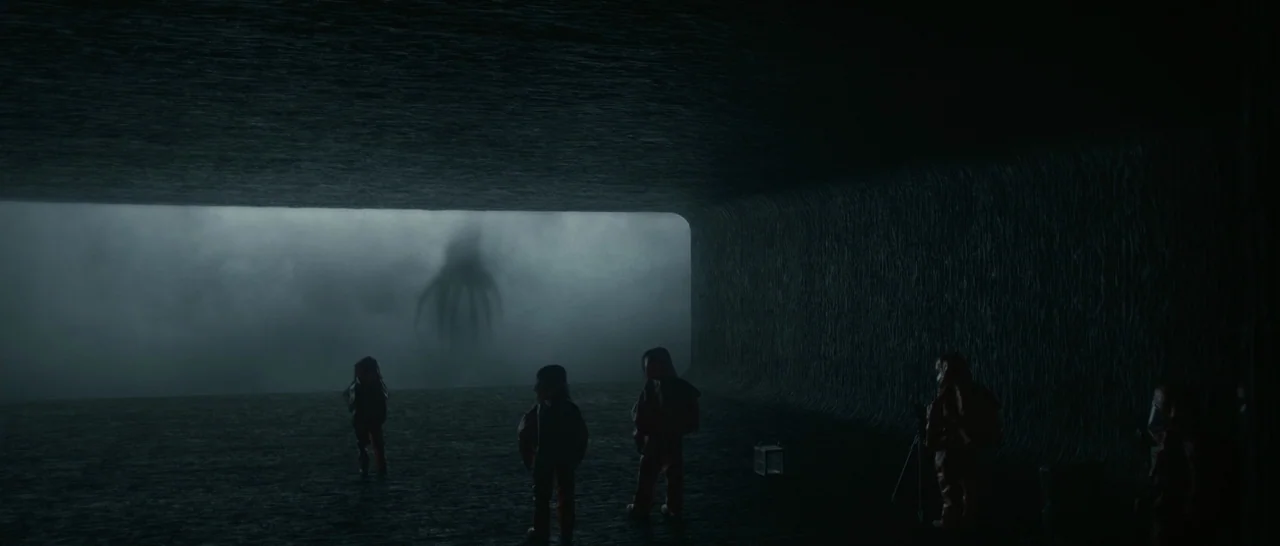 Arrival (2016) movie still: wide — A dark, smooth-walled stone chamber opens to a vast, misty landscape. Five figures in jumpsuits observe a giant…