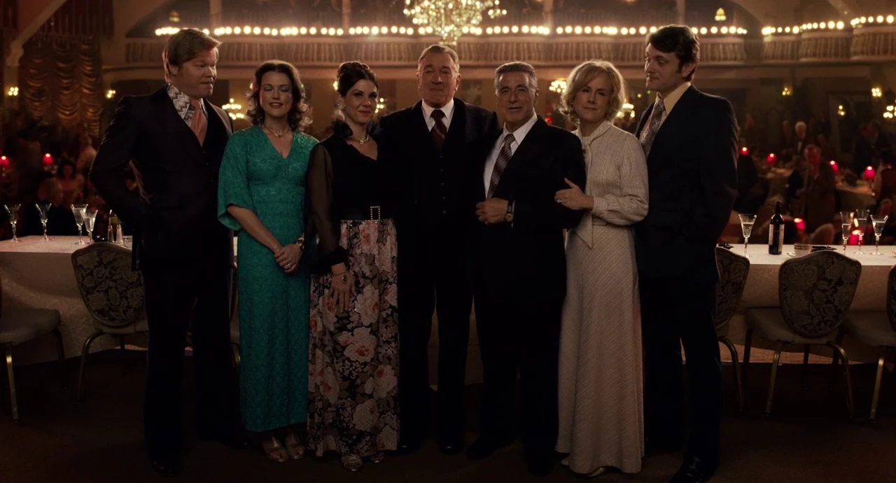 The Irishman (2019) movie still: fullbody — A group of seven people pose for a formal portrait in a large ballroom. The room features long banquet…