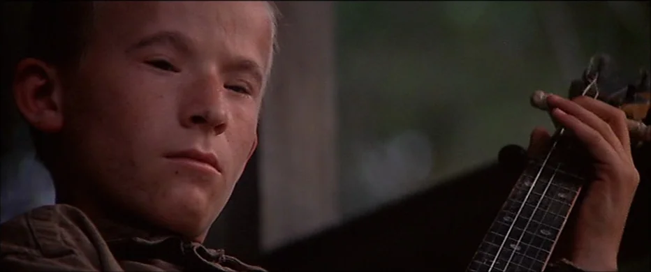 Deliverance (1972) movie still: closeup — A rural setting with a boy holding a stringed instrument. He stands outdoors while playing a banjo with…