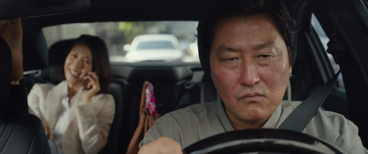 Parasite (2019) movie still: medium — Inside a modern vehicle, a man drives while a passenger sits behind him. The passenger is talking on a…