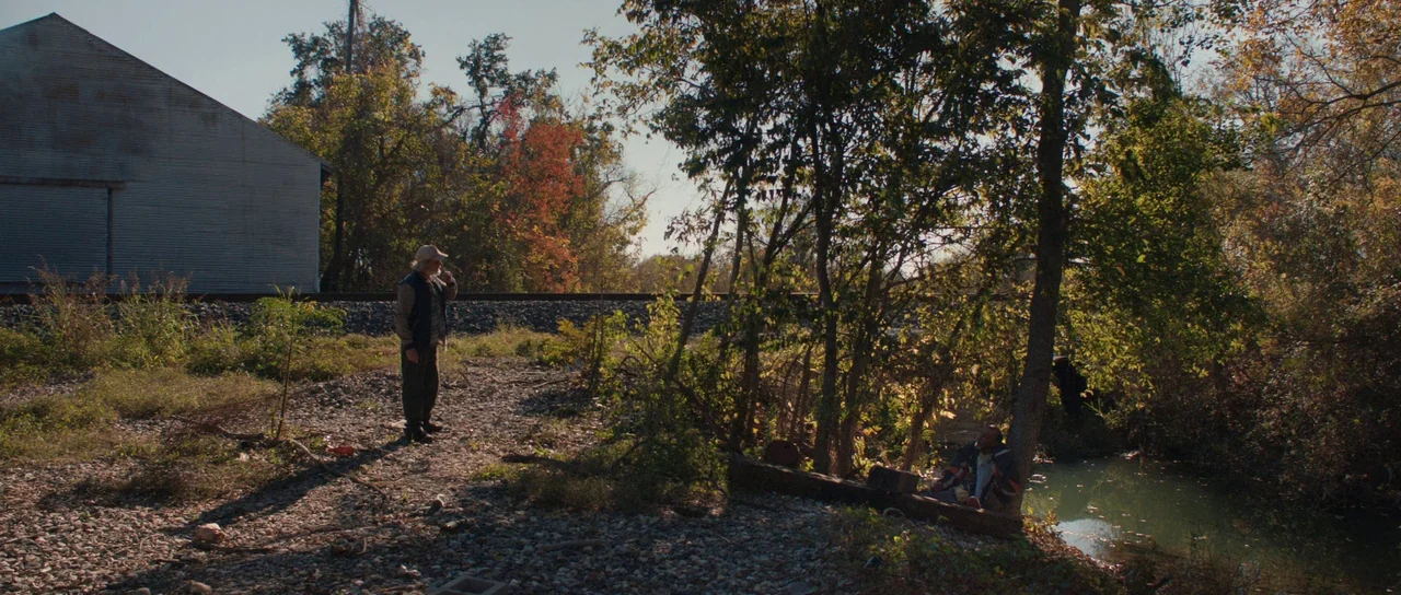 Joe (2013) movie still: wide — A gravel area beside railroad tracks features an industrial shed and autumn trees nearby. One man stands while…