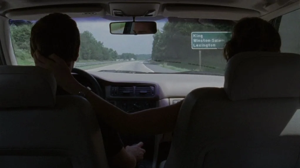 Junebug (2005) movie still: medium — A view from the back seat of a car traveling down a highway. The passenger reaches forward to touch the driver…
