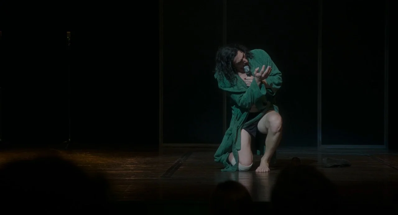Annette (2021) movie still: medium — A dark wooden platform serves as a bare stage in a dimly lit hall. A man kneels in the center of the pool of…