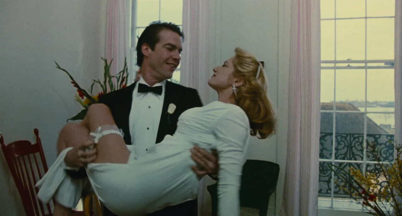 The Big Easy (1986) movie still: medium — A sunlit room features floor-to-ceiling windows with sheer curtains and floral arrangements. A man in…