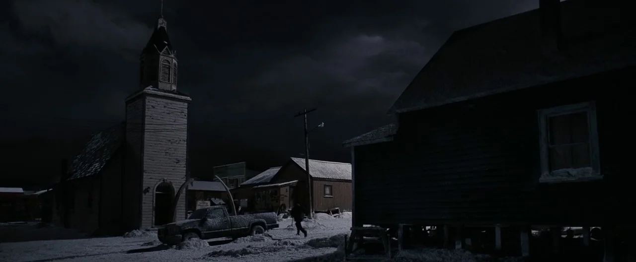 30 Days of Night (2007) movie still: wide — A desolate town square sits under a dark night sky covered in heavy snow. A church steeple and a few…