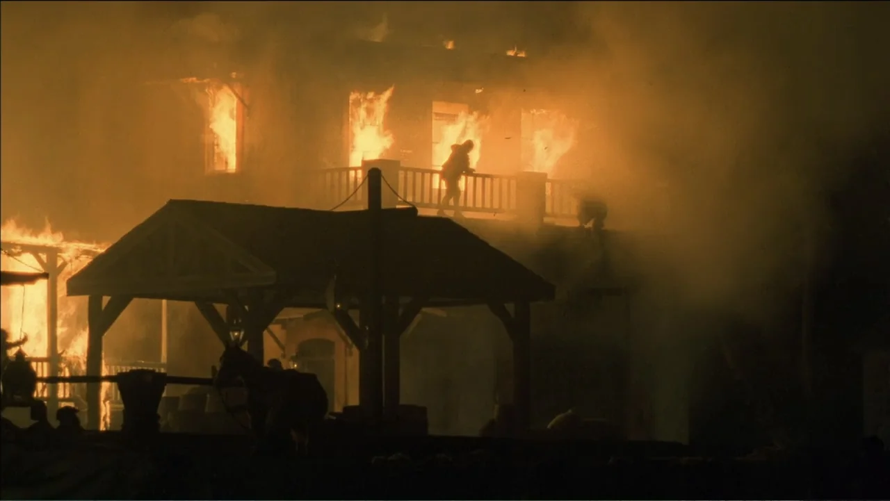 Empire Queen: The Golden Age of Magic (2024) movie still: wide — A two-story wooden structure is engulfed in large flames amidst thick billows of…