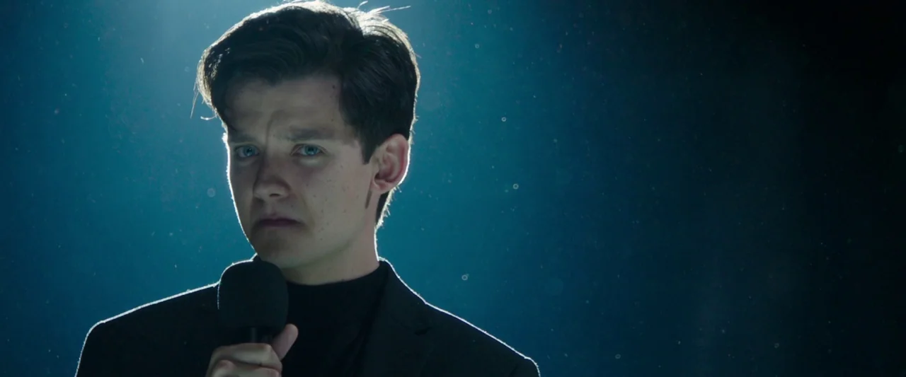 Greed (2019) movie still: closeup — A dark environment with visible dust particles drifting through a narrow spotlight beam. A young man stands…