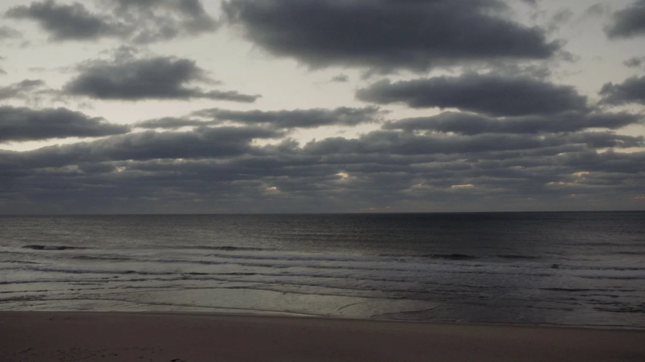 Marjorie Prime (2017) movie still: establishing — An empty beach sits before a dark, overcast ocean under a heavily clouded sky. Low waves roll onto…