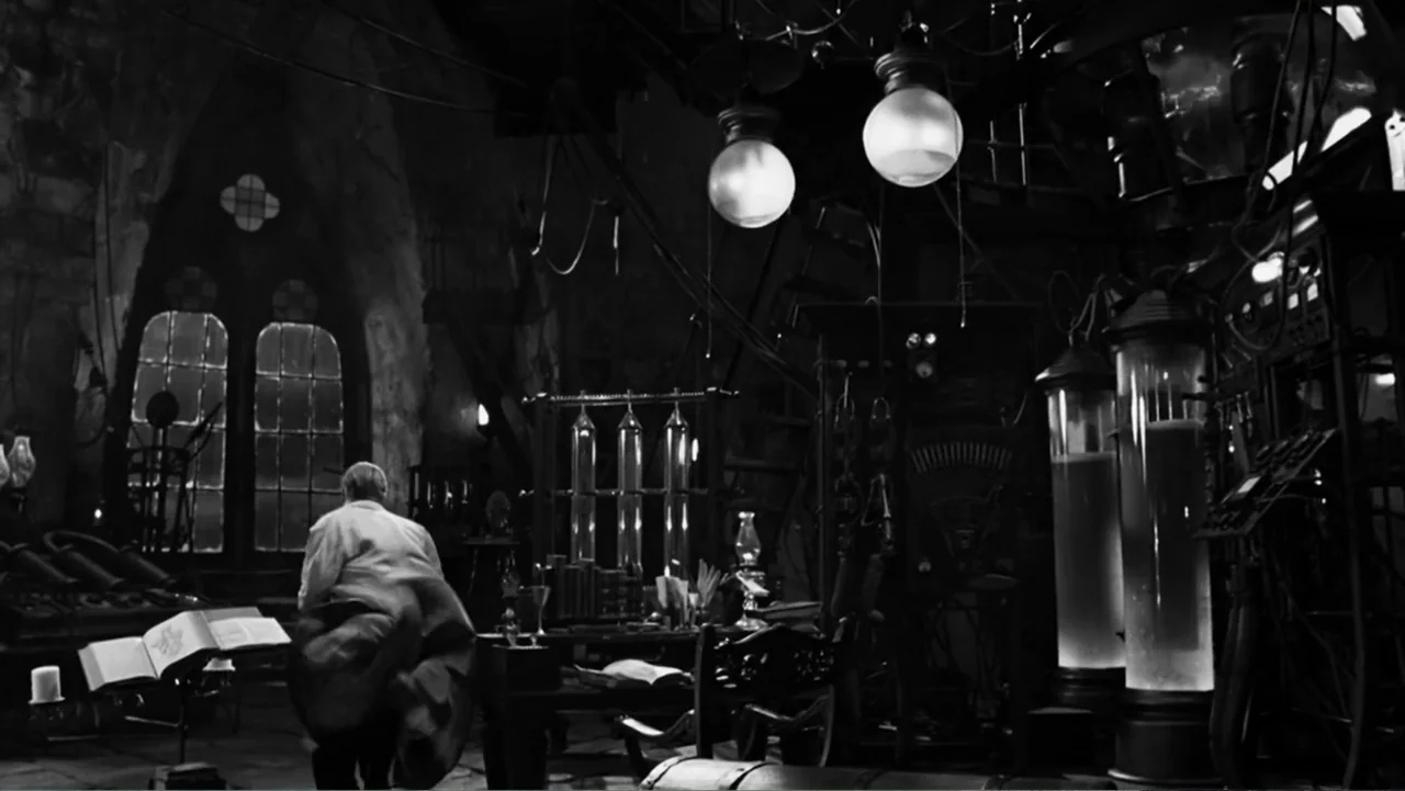 Van Helsing (2004) movie still: wide — A stone-walled laboratory featuring large glass tubes, electrical coils, and gothic arched windows. A man in…