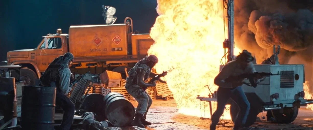 True Lies (1994) movie still: wide — An industrial fueling area filled with metal barrels and heavy machinery sits under night. Three armed figures…