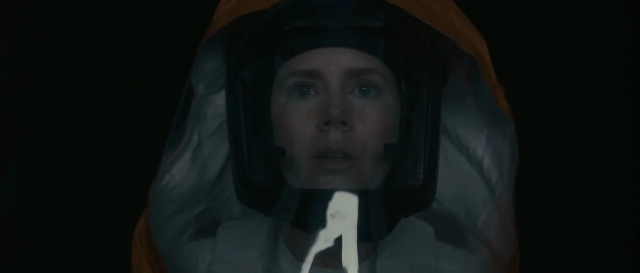 Arrival (2016) movie still: closeup — A dark, void-like environment surrounds a person wearing a protective orange hood and helmet. The individual…