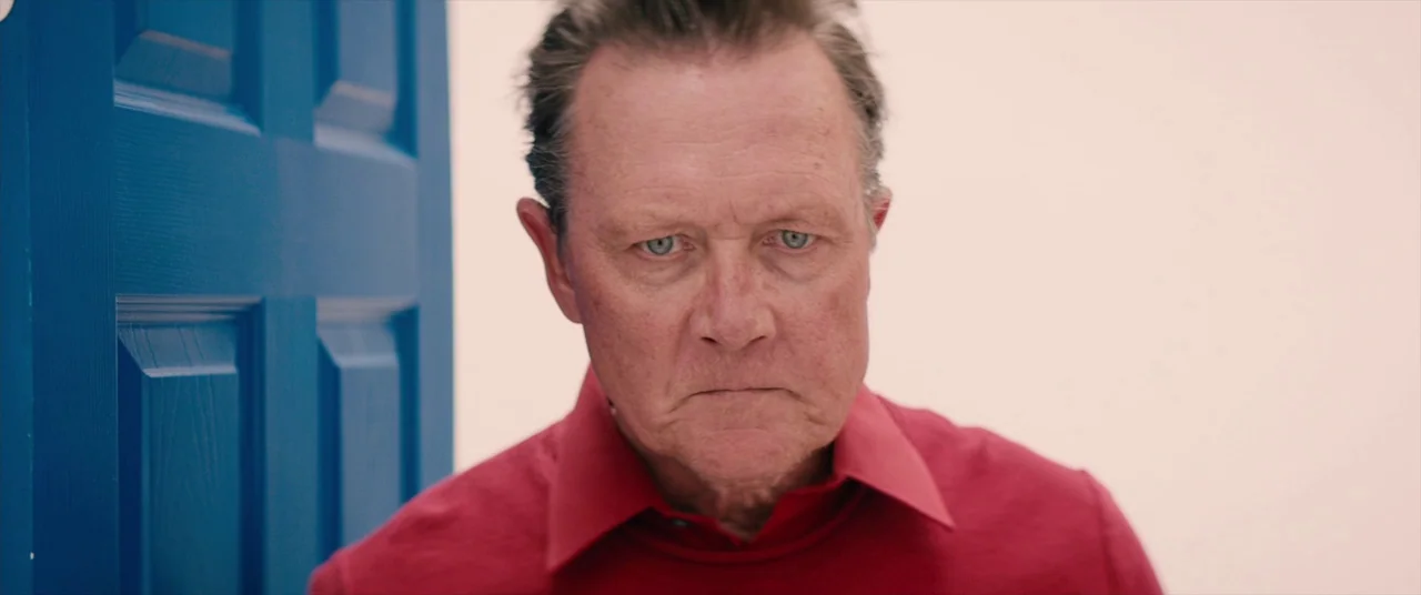 Tone Deaf (2019) movie still: closeup — A man stands in a brightly lit, sterile room with a solid blue door. He stares directly ahead into the lens…