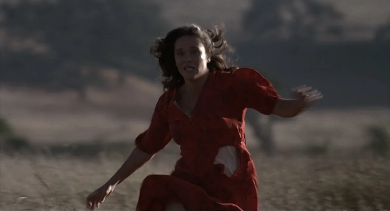 Of Mice and Men (1992) movie still: medium — A dry, grassy field stretches into the background under a late afternoon sun. A woman in a torn red…