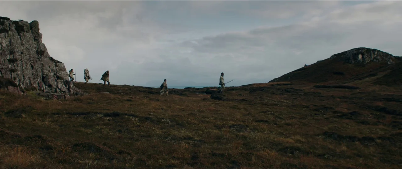 Out of Darkness (2022) movie still: wide — A vast, uneven highland moor extends toward a distant sea under a cloudy sky. Five individuals walk…