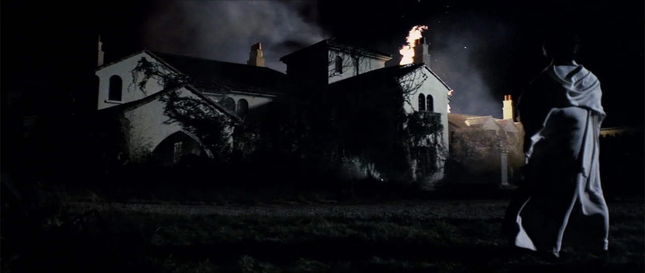 Elektra (2005) movie still: wide — A large multi-story stucco mansion burns at night with smoke rising into the air. A person viewed from behind in…