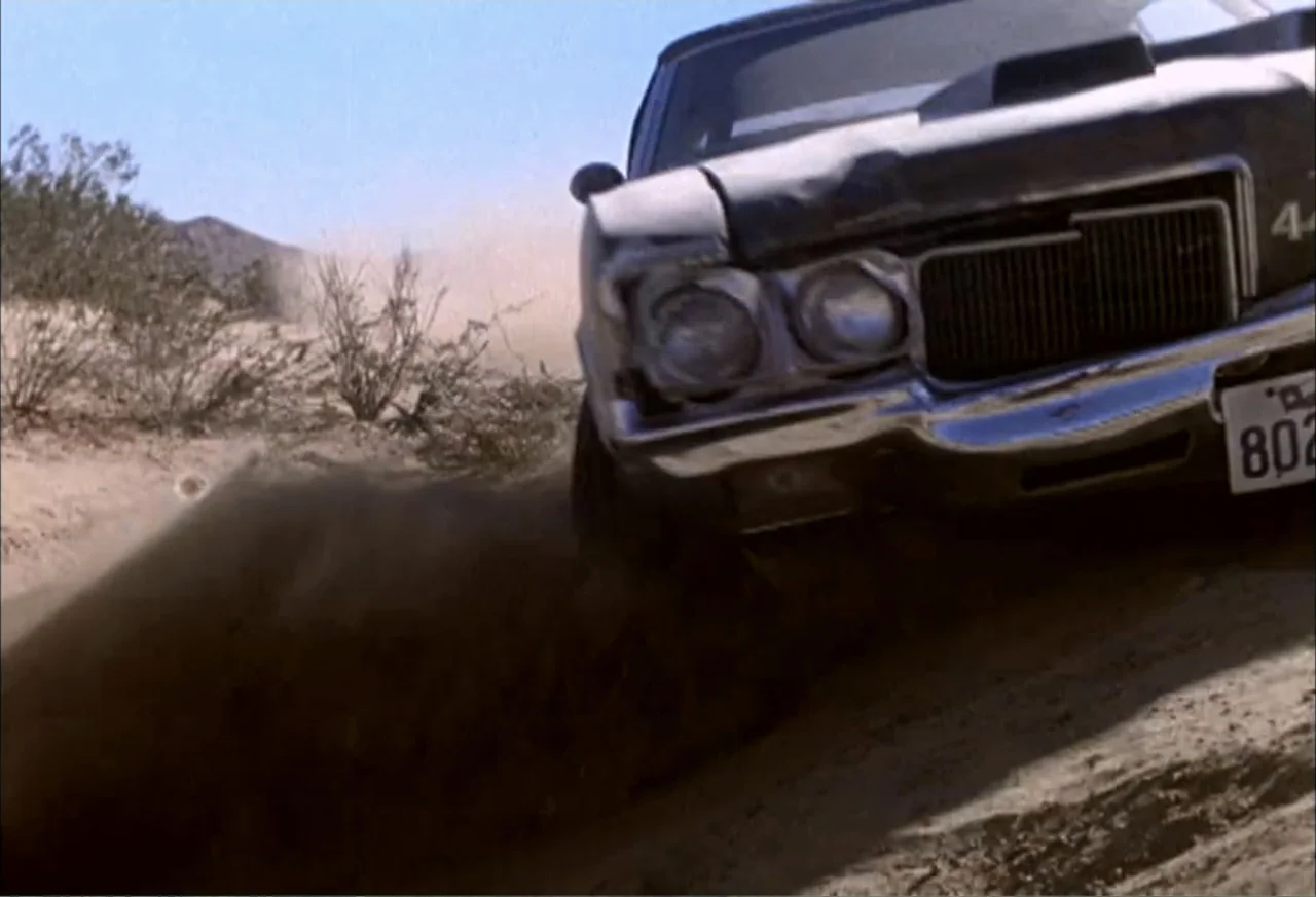Black Cat Run (1998) movie still: medium — A vintage sedan drives along a dry dirt path in a vast desert landscape. The car kicks up a cloud of dust…