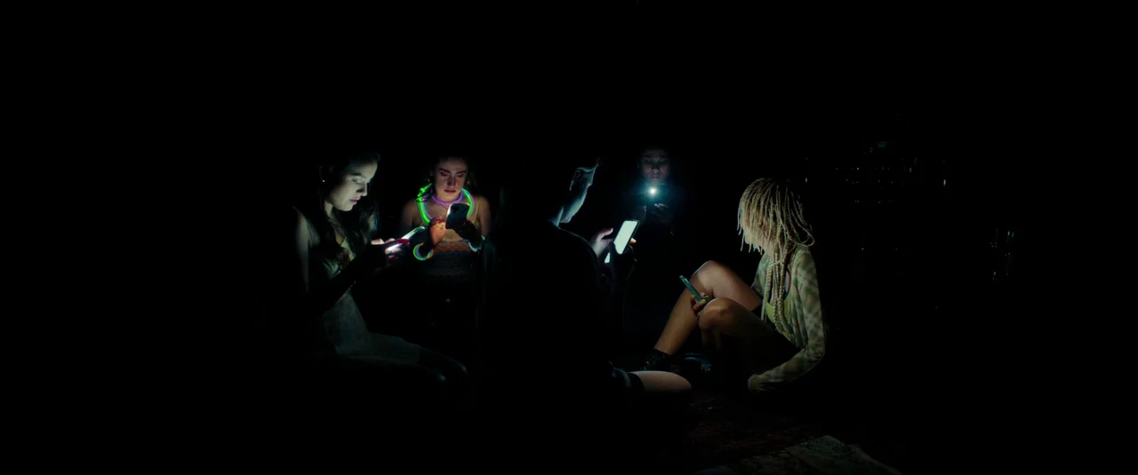 Bodies Bodies Bodies (2022) movie still: wide — A group of five young people sit in a completely dark room. They are looking down at their…