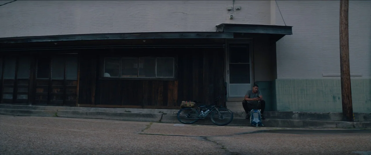 Rebel Ridge (2024) movie still: wide — A quiet urban storefront features a weathered wooden facade and a pale concrete curb. A man sits alone on the…