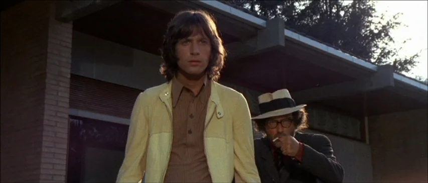 Four Flies On Grey Velvet (1971) movie still: medium — A modern building with brick masonry and horizontal roof eaves forms the backdrop. A young…