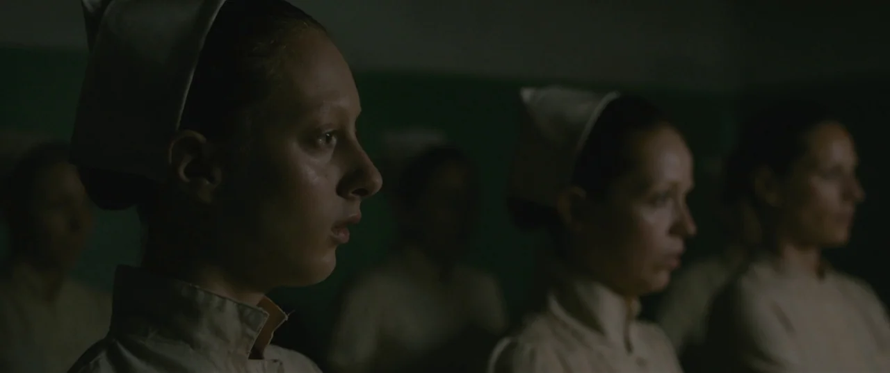 Evolution (2015) movie still: medium — A dark, dimly lit room with a muted green wall in the background. A group of nurses in uniform stand in a…