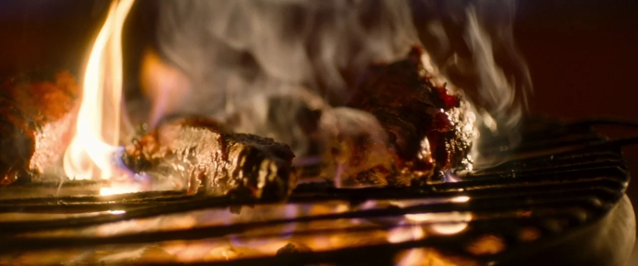Baskin (2015) movie still: closeup — Several cuts of raw meat sear on a hot metal barbecue grill at night. Visible flames and smoke emanate from…