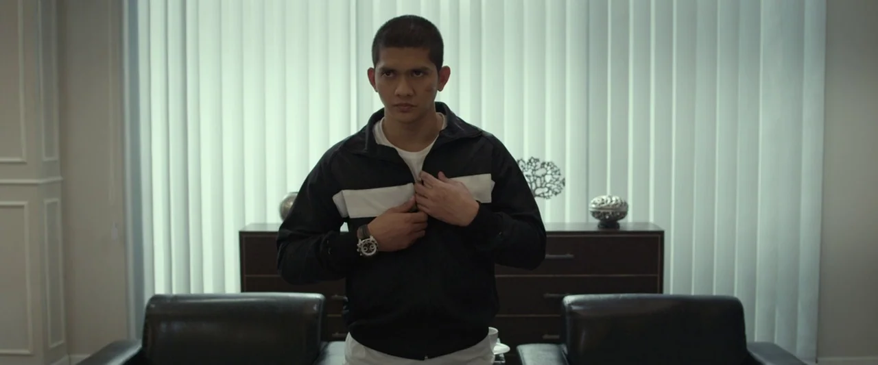 The Raid 2 (2014) movie still: medium — A man stands in a modern room with vertical blinds and dark wooden furniture. He pulls the zipper of his…