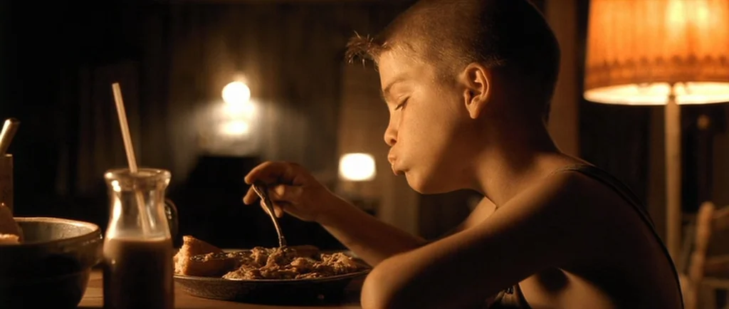 O Brother, Where Art Thou? (2000) movie still: medium — A dark, dimly lit indoor space features a young boy eating dinner alone. A warm lamp…