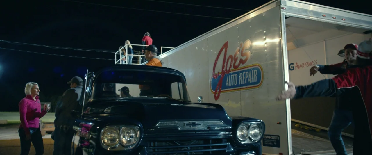 God Bless the Broken Road (2018) movie still: wide — A nighttime scene in a paved lot featuring a dark classic pickup truck. Several people gather…